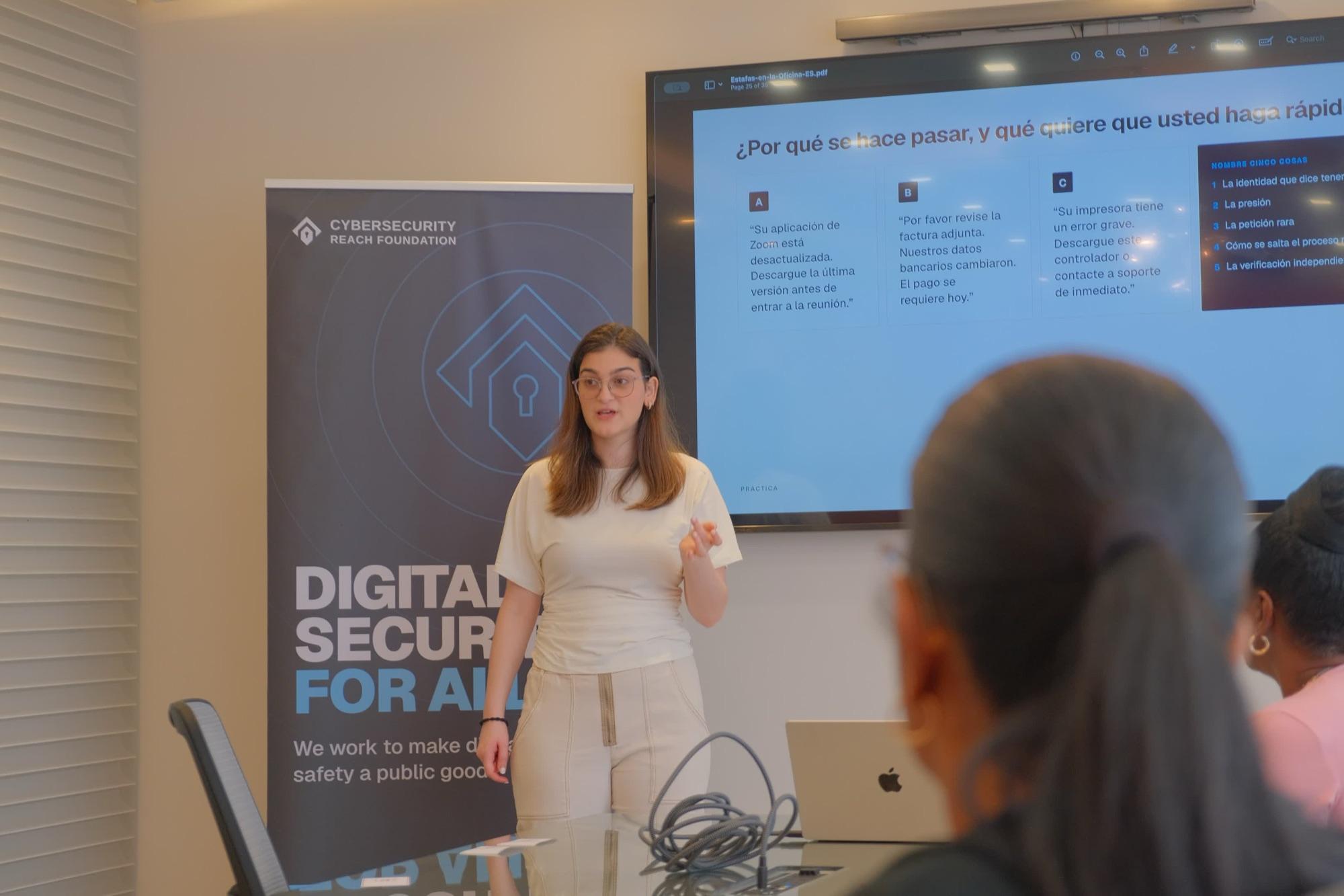 A volunteer speaking to seated staff in an office meeting room, with a Spanish-language scam awareness slide on the screen behind her