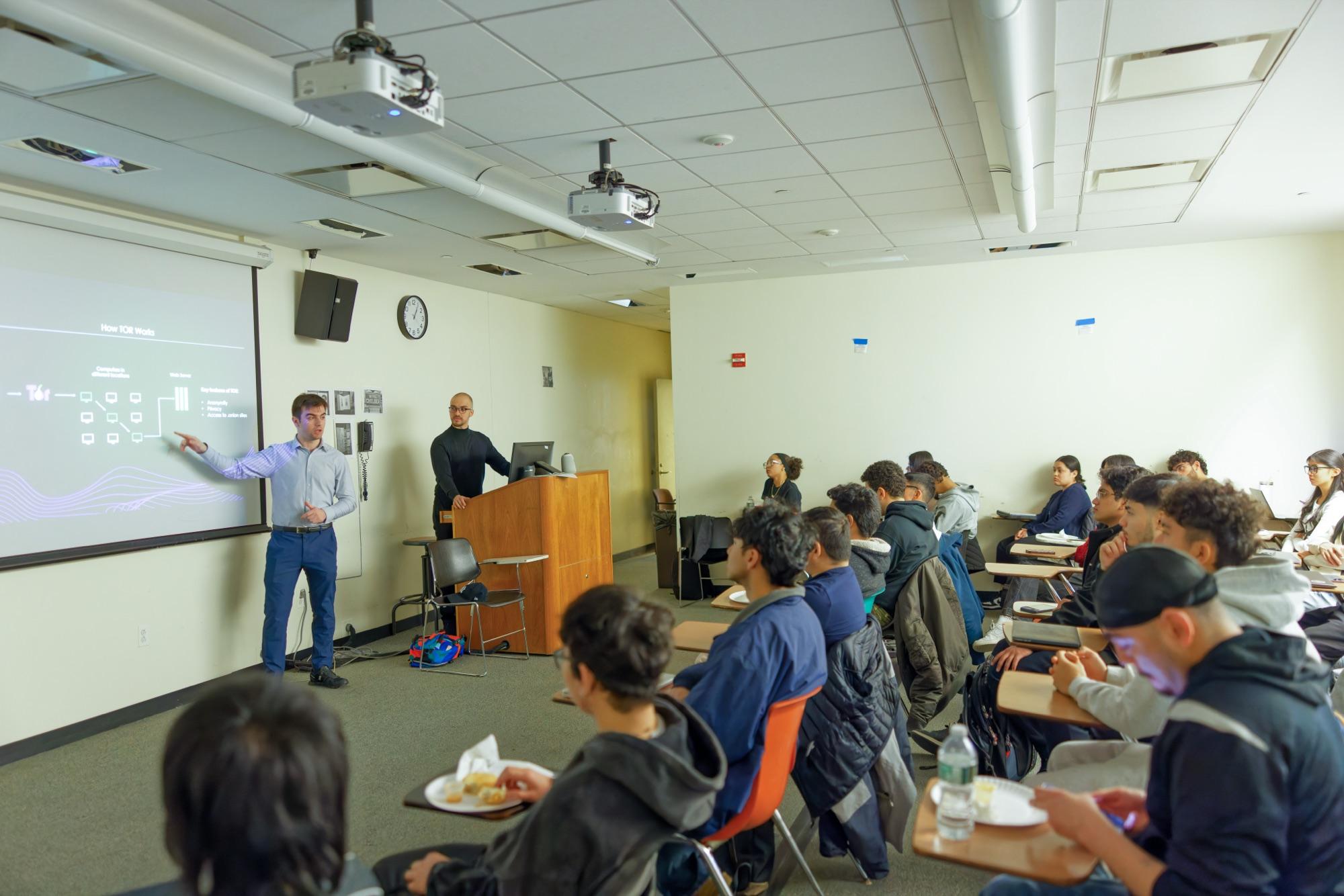 A Cybersecurity Reach Foundation volunteer points at a slide explaining how Tor works, in front of a full classroom of university students