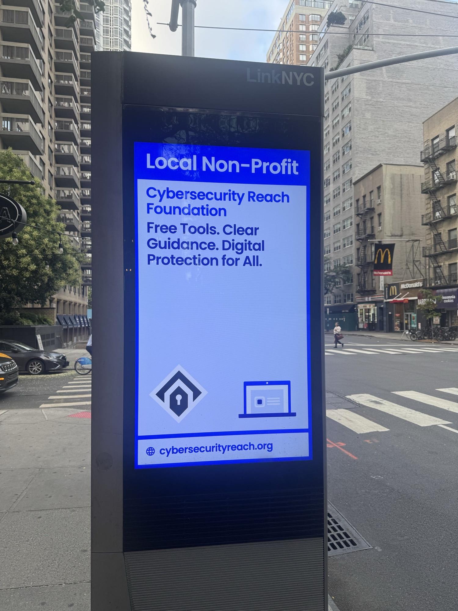 A LinkNYC street kiosk on a Manhattan corner displaying a Cybersecurity Reach Foundation advert reading Free Tools, Clear Guidance, Digital Protection for All